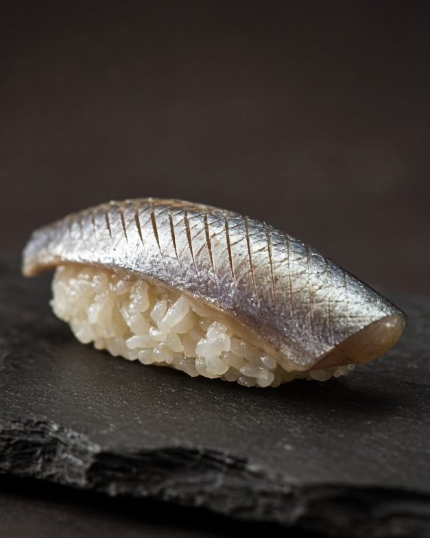One piece of silver-skinned nigiri on dark slate, with a little wasabi